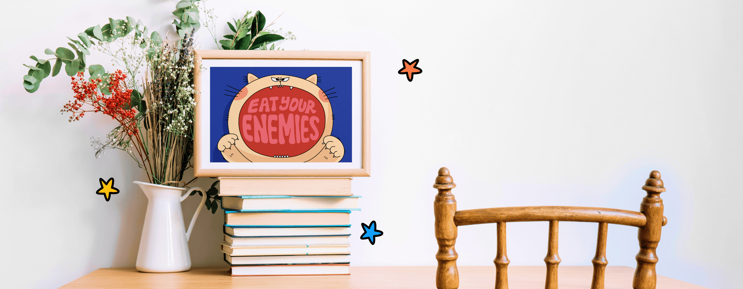 A wooden framed art print showing a cat saying eat your enemies humorously, placed on a stack of books with a plant and chair nearby, against a white background