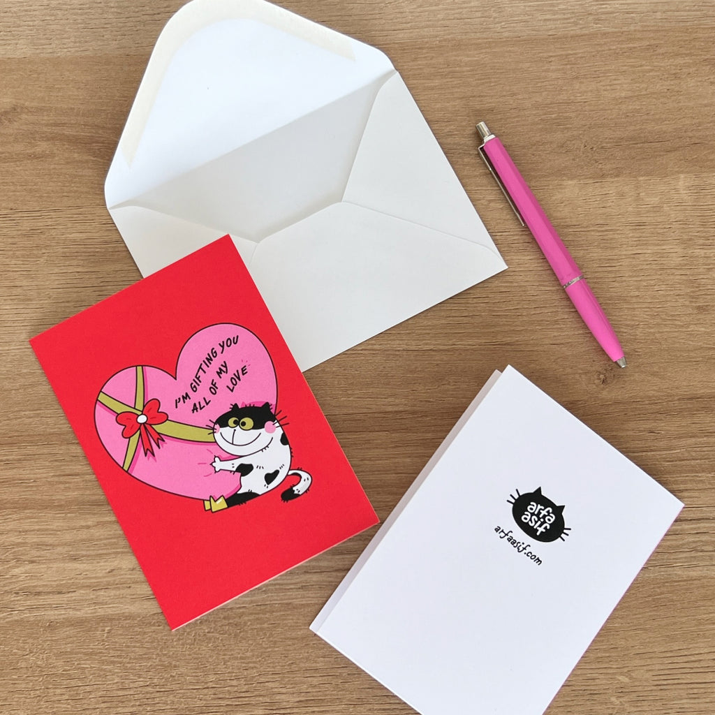 Red card with cartoon cat holding a heart, white envelope, pink pen, and white card with black logo on a wooden surface.