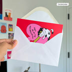 Hand holding a white envelope with a pink card featuring a cartoon cat and text.