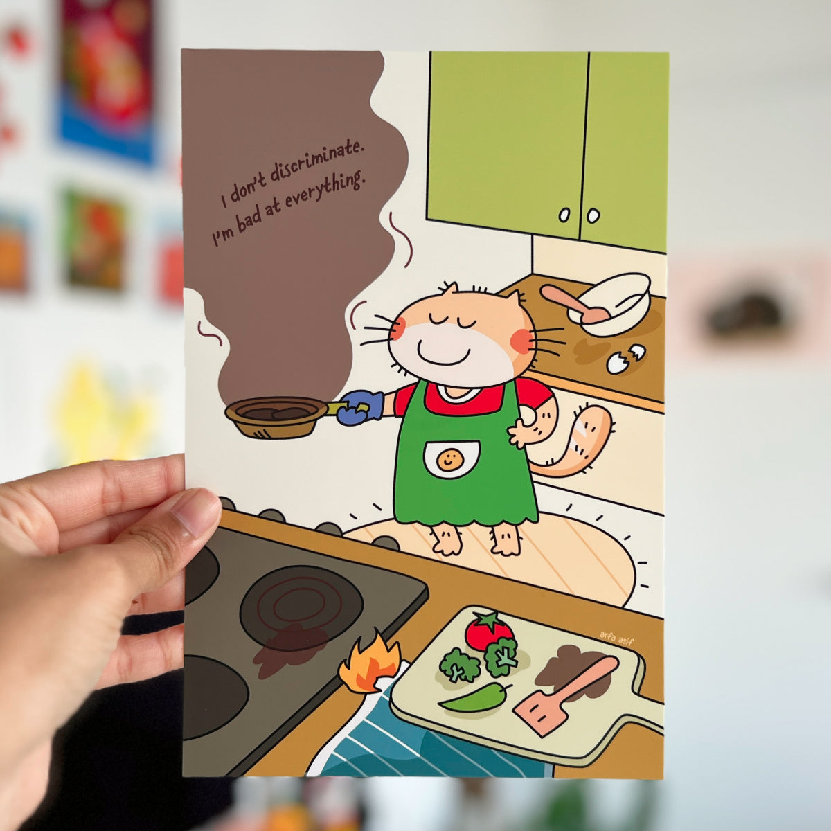 Cartoon character in a kitchen holding a frying pan with text, held by a hand against a blurred background.