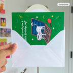 Hand holding a green birthday card with a cat illustration and envelope.