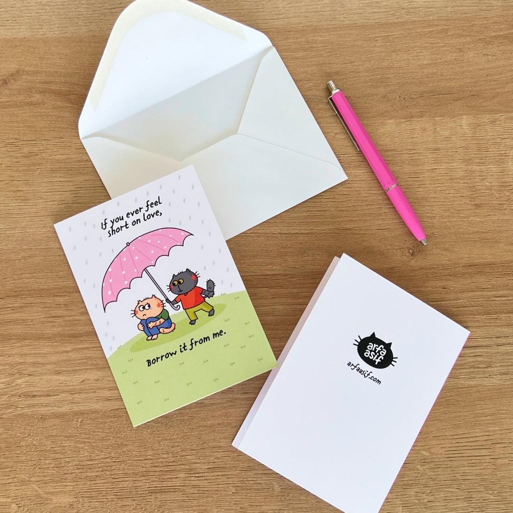 Greeting card with cat characters and text on a wooden surface with an envelope and pen.