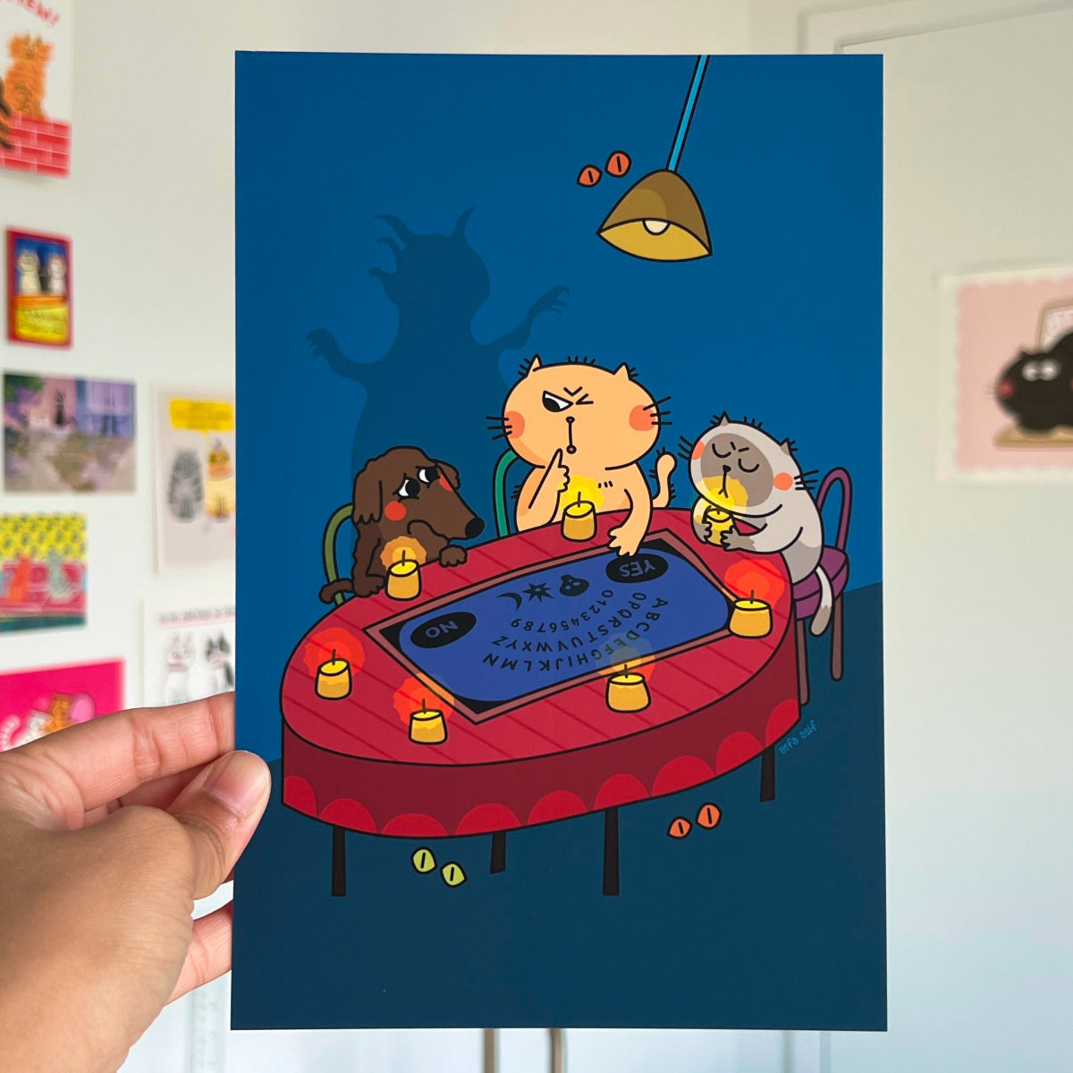 Cartoon cats playing with Ouija Board on a table with a hand holding the poster.