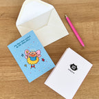 Greeting card with cartoon cat illustration on a wooden surface with an envelope and pen.