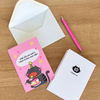 Pink greeting card with a cat illustration and white envelope on a wooden surface.