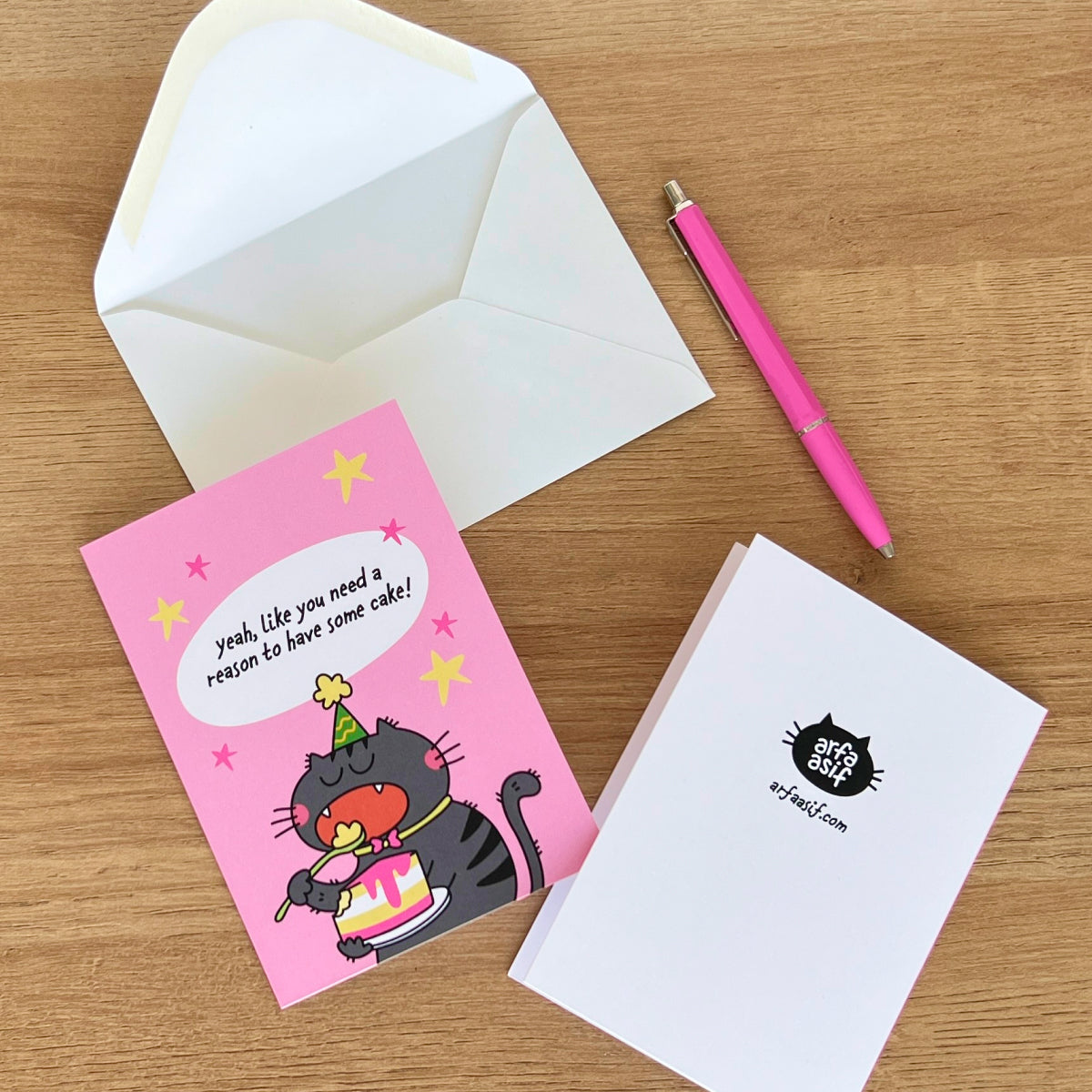 Pink greeting card with a cat illustration and white envelope on a wooden surface.