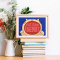 Framed artwork with a cat illustration and text 'Eat Your Enemies' on a stack of books, surrounded by plants.