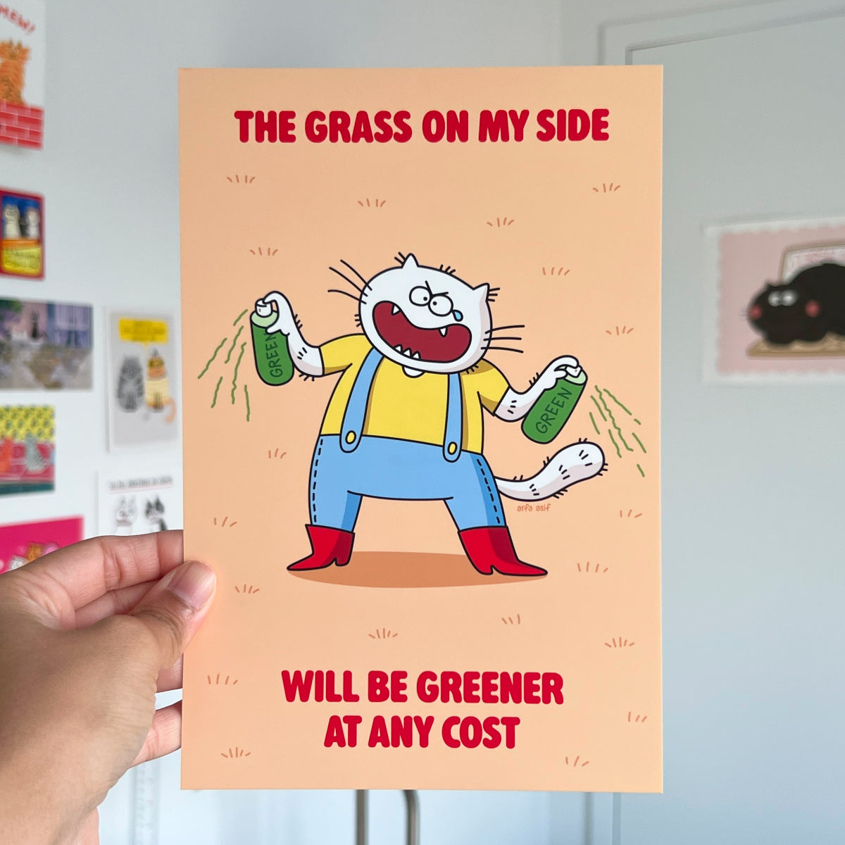 Art print with a cat character holding grass and text on an orange background, held by a hand.