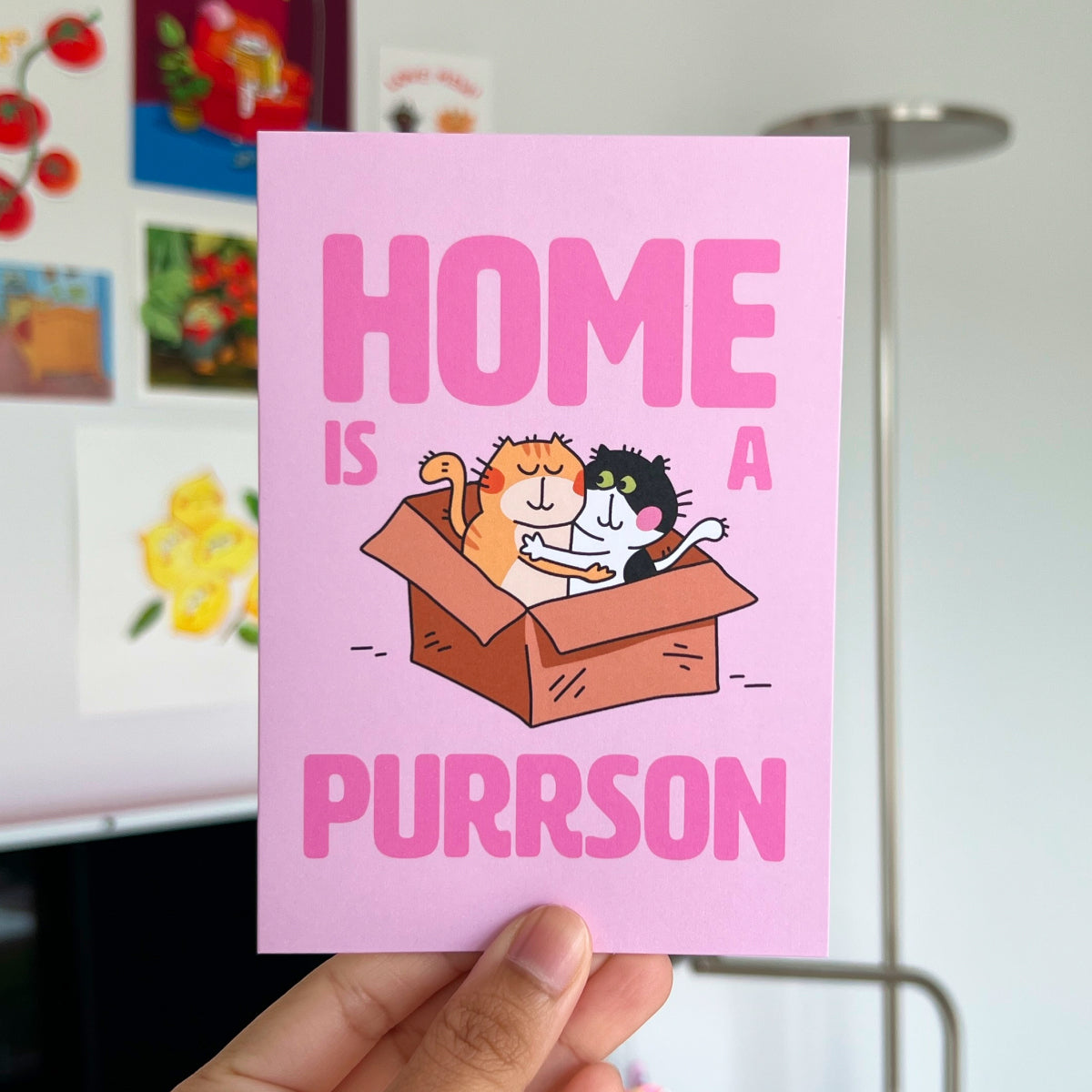 Card with cats and text 'Home is a purrson' held by a hand in front of a colorful background.