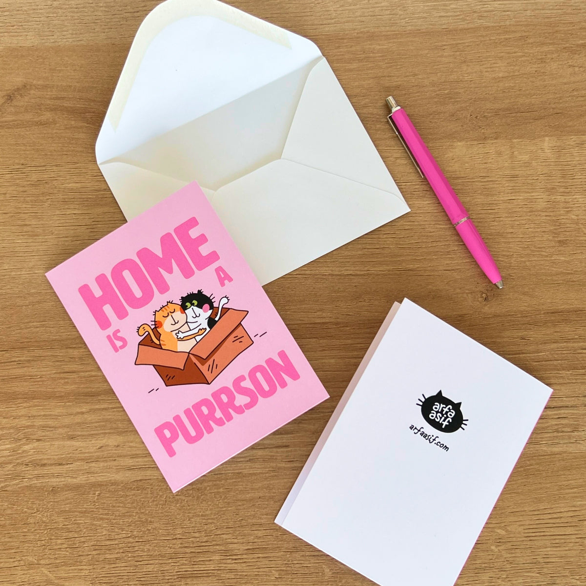 Pink greeting card with cat illustration and text, white envelope, pink pen on wooden surface