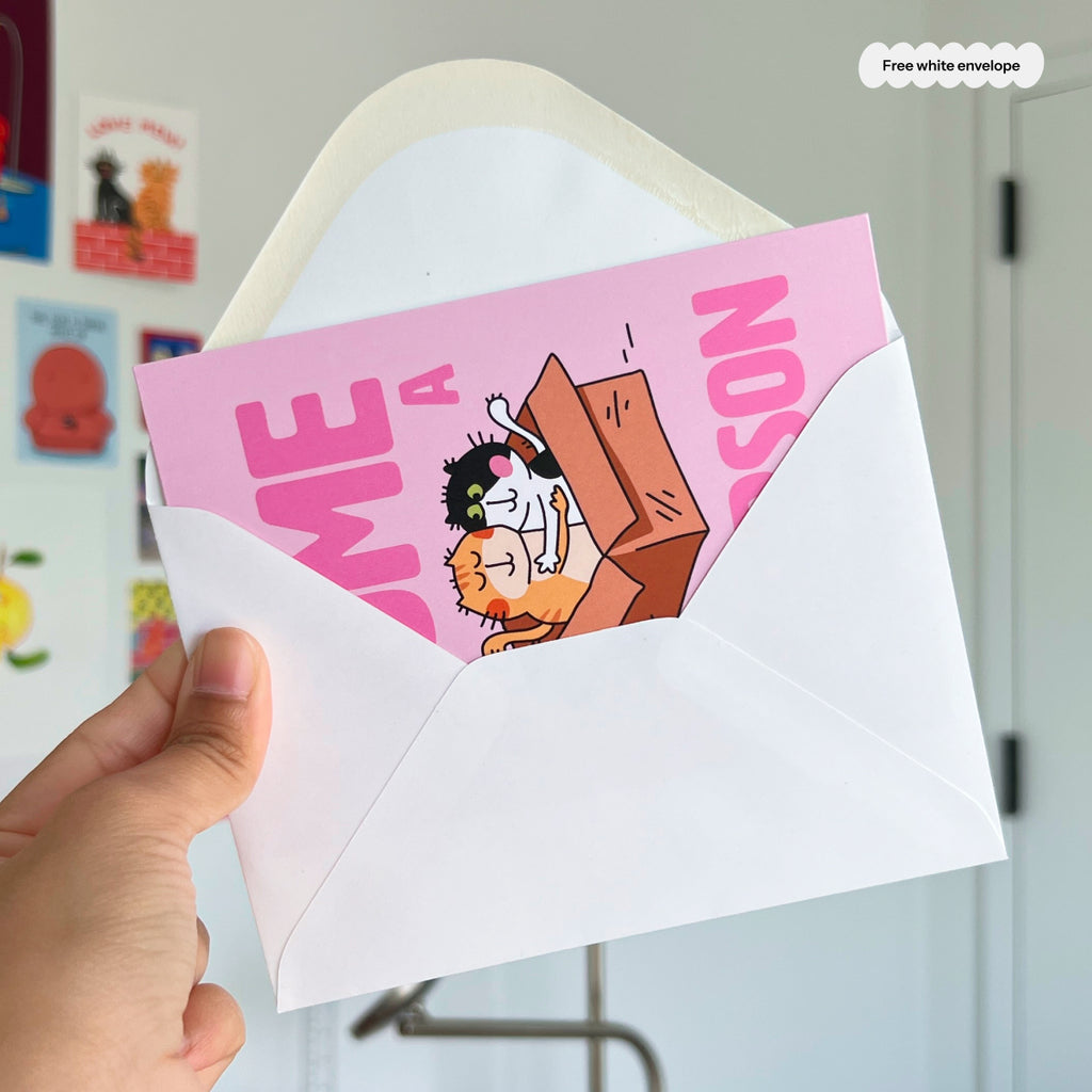 Hand holding a pink card with cartoon character and envelope in front of a white wall.