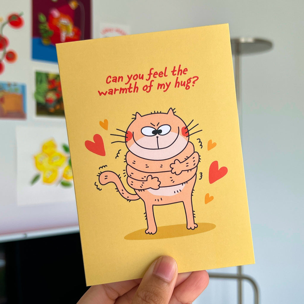 Card with a cartoon cat hugging itself and text 'Can you feel the warmth of my hug?' held by a hand.