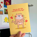 Card with a cartoon cat hugging itself and text 'Can you feel the warmth of my hug?' held by a hand.