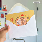Hand holding a white envelope with a cartoon card inside, featuring a character and text.