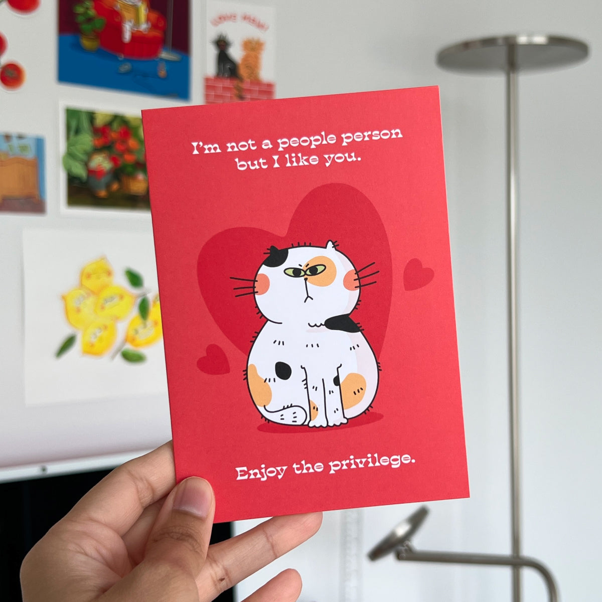 Red card with a cat illustration and text held by a hand, with a blurred background
