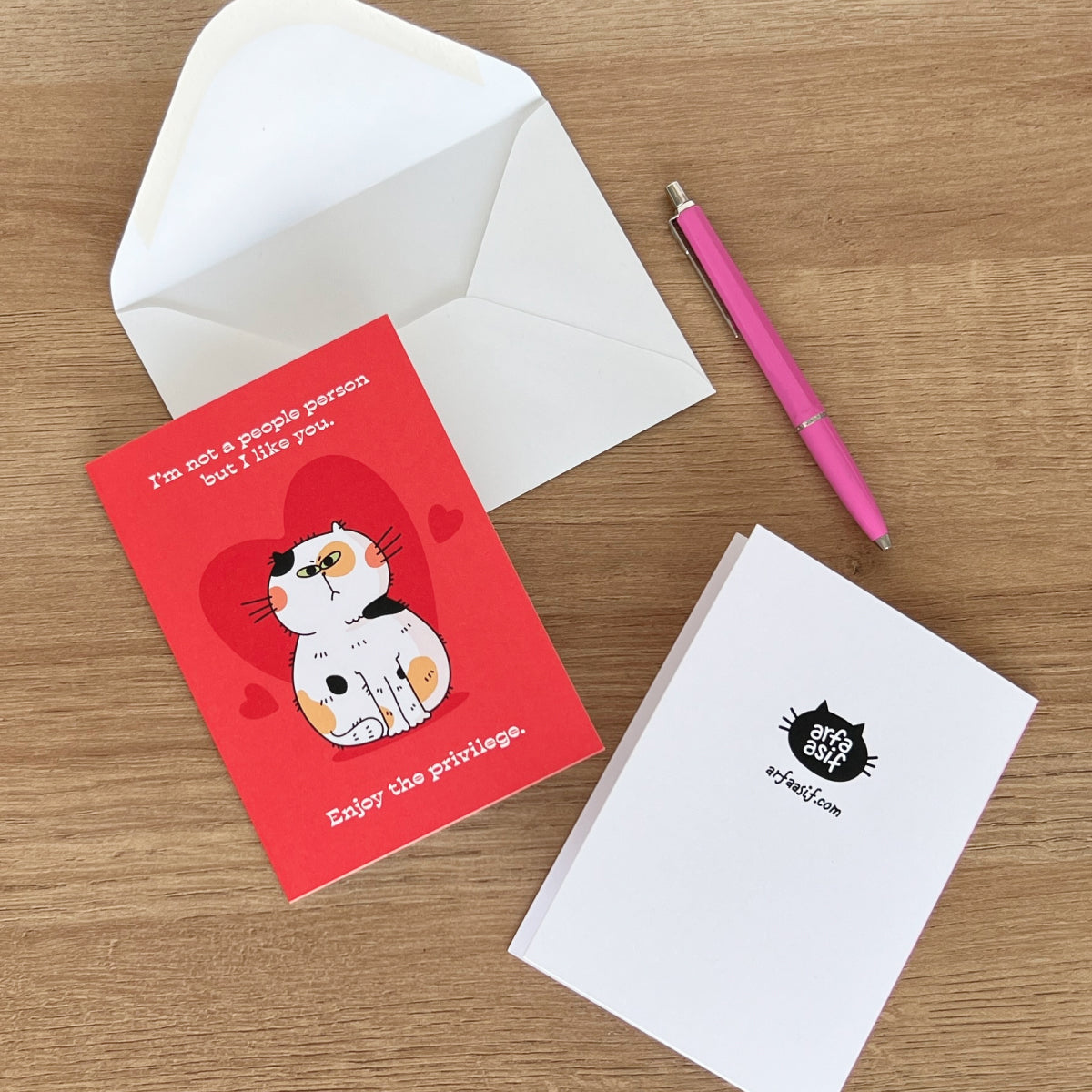 Card with cat illustration and text, white envelope, pink pen, and white card with black cat logo on a wooden surface.