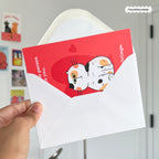 Red card with cartoon cat and white envelope held by a hand against a light gray wall.