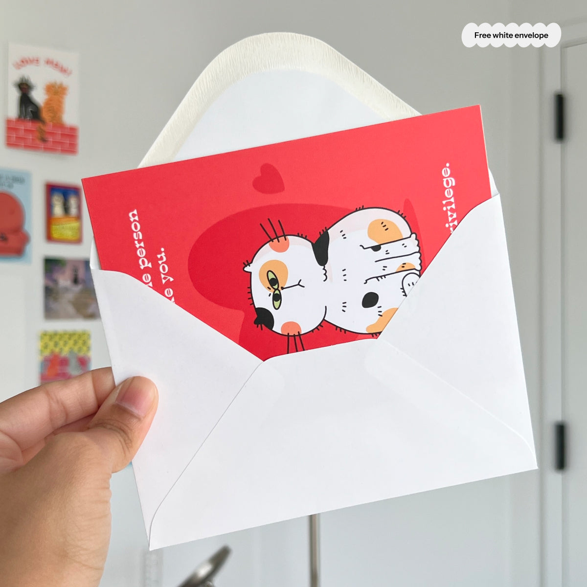 Red card with cartoon cat and white envelope held by a hand against a light gray wall.