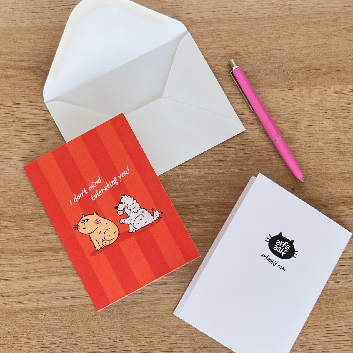 Red greeting card with cartoon dog and cat, and white envelope on a wooden surface with a pink pen.