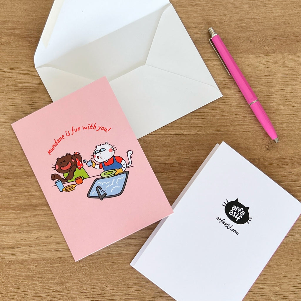 Pink greeting card with cat characters and text, white envelope, pink pen on wooden surface