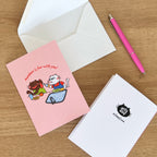 Pink greeting card with cat characters and text, white envelope, pink pen on wooden surface