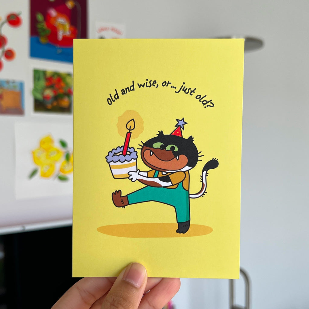 Greeting card with cartoon cat holding a cake and a candle, text 'old and wise, or... just old?' held by a hand.