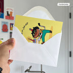 Hand holding a card with a cat character and text, inside a white envelope.