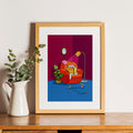 Framed artwork of a cat on an armchair with a plant and clock on a wooden surface.