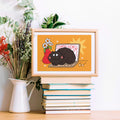 Framed illustration of a black cat on a laptop with flowers, placed on books with plants in the background.