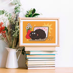 Framed illustration of a black cat on a laptop with flowers, placed on books with plants in the background.