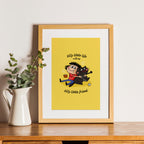 Framed cartoon print with a yellow background featuring a character and text, placed on a wooden surface with a plant beside it.