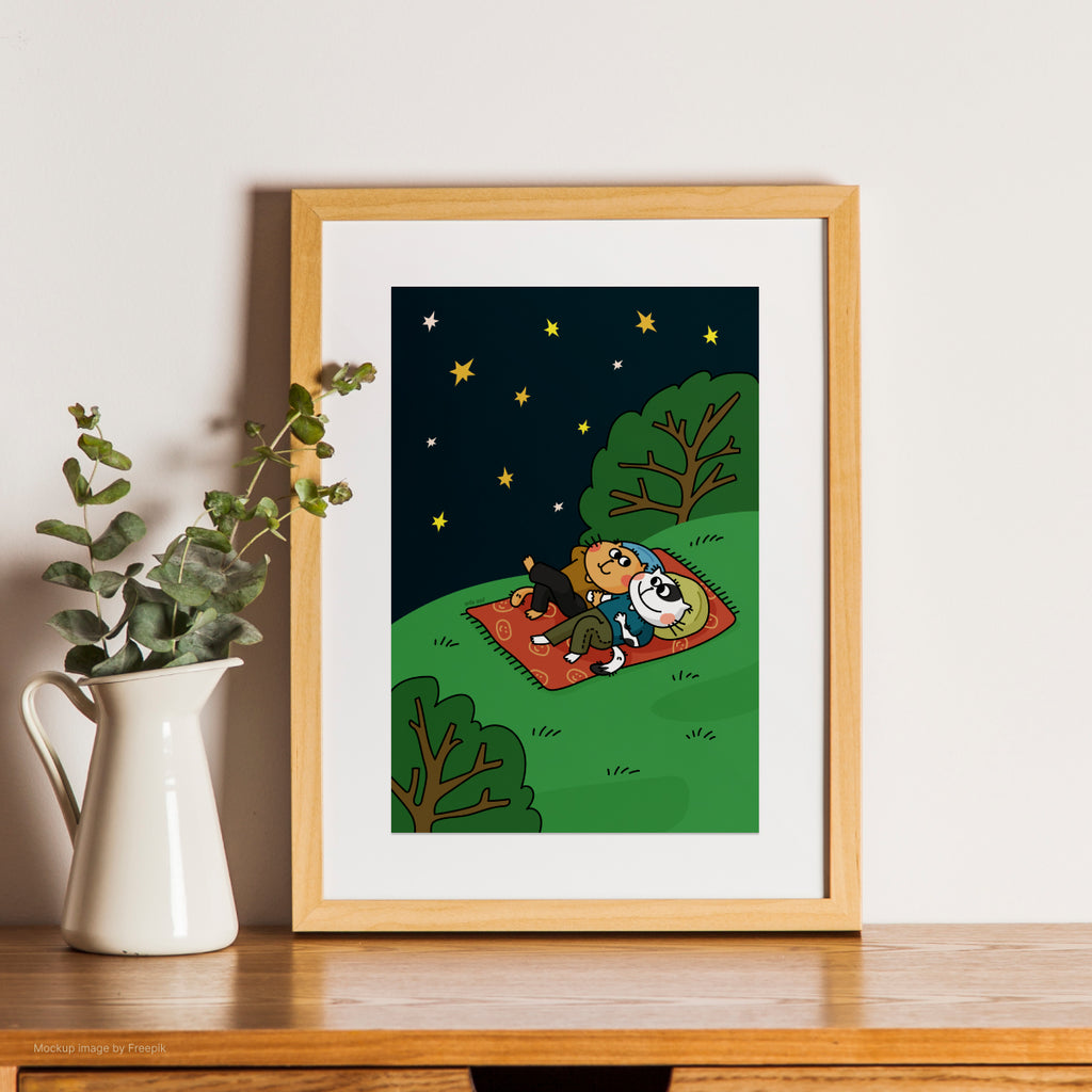 Framed illustration of two cats on a blanket under stars, placed on a wooden surface with a plant beside it.