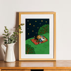 Framed illustration of two cats on a blanket under stars, placed on a wooden surface with a plant beside it.