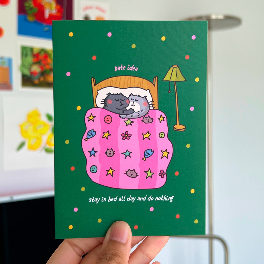 Card with cartoon cats under a pink blanket on a green background, held by a hand.