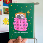 Card with cartoon cats under a pink blanket on a green background, held by a hand.