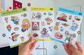 Three cat sticker sheets held by hands with a colorful background