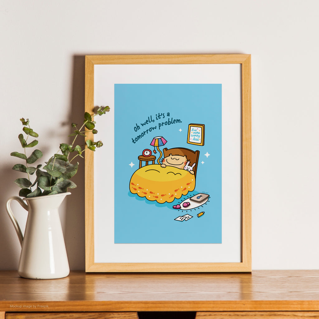 Framed cartoon print of a cat in bed with a blue sky background, sitting on a wooden surface next to a white pitcher with greenery.