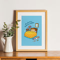 Framed cartoon print of a cat in bed with a blue sky background, sitting on a wooden surface next to a white pitcher with greenery.