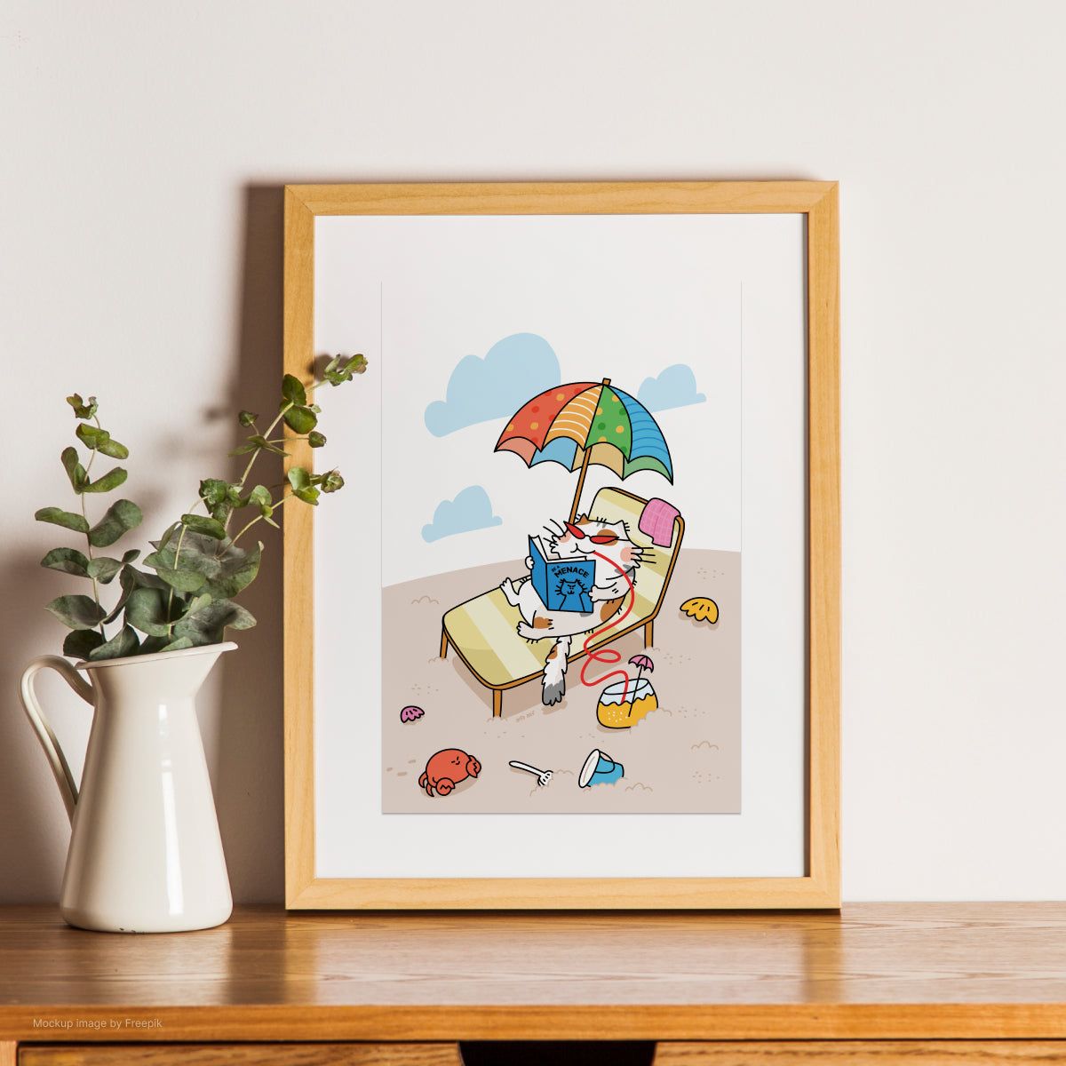 Framed illustration of a cat reading a book under an umbrella on a beach, placed on a wooden surface with a plant beside it.