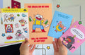 Four colorful paper goods held by a person against a white wall.