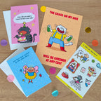 One art print, two colorful greeting cards and a sticker sheet on a wooden surface with decorative confetti.