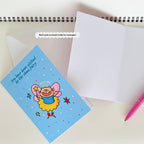 Greeting card with funny cat fairy design on a white surface with an envelope and pen.