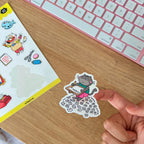 A sticker of a cartoon cat sitting on a pile of papers, held by a hand against a wooden background.